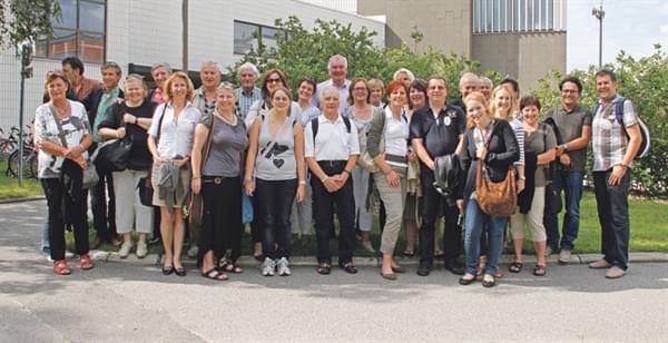 Eine Gruppe von etwa dreißig Erwachsenen posiert lächelnd für ein Gruppenfoto im Freien, vor modernen Gebäuden und grüner Vegetation.