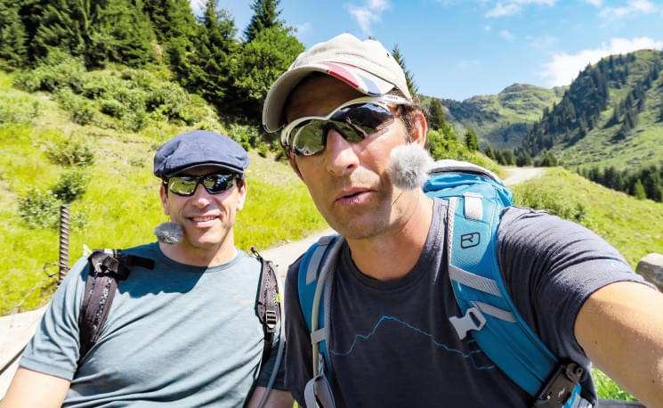Zwei Männer posieren beim Wandern in den Bergen. Beide tragen Sonnenbrillen und Mützen, umgeben von grüner Landschaft und klarem Himmel im Hintergrund.