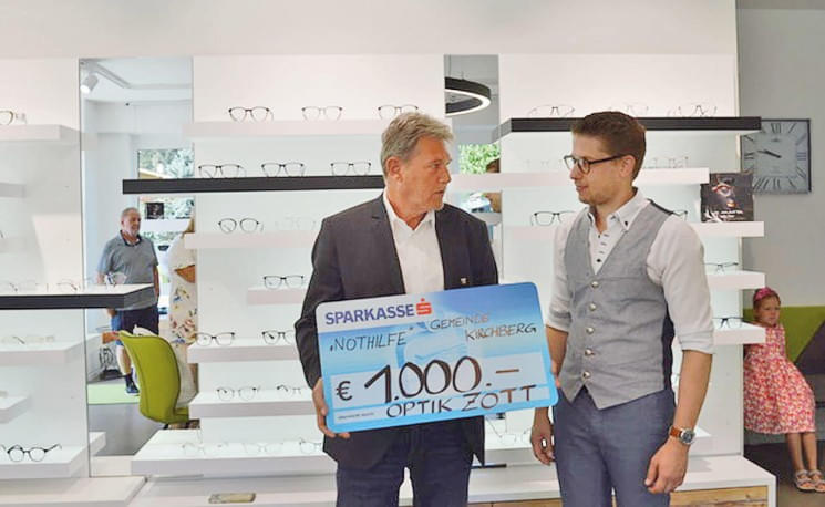 Zwei Männer stehen in einem Optikergeschäft und halten einen großen Scheck über 1000 Euro. Im Hintergrund sind Brillenregale zu sehen.
