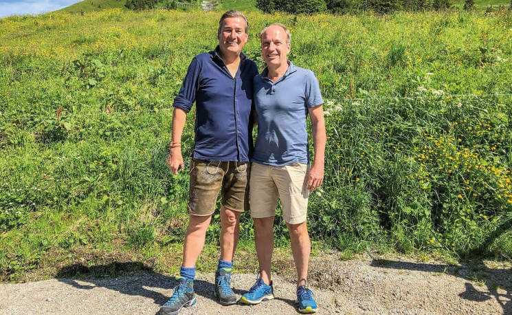 Zwei Männer stehen lächelnd Arm in Arm vor einer grünen Wiese mit blühenden Pflanzen. Beide tragen Freizeitkleidung und wandertaugliche Schuhe.