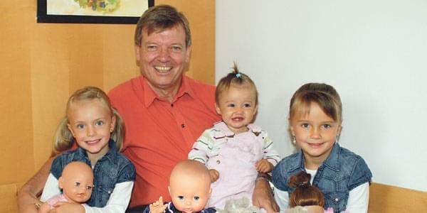Ein älterer Mann sitzt lächelnd inmitten von drei jungen Mädchen. Sie halten Puppen in den Händen und scheinen fröhlich zu sein.