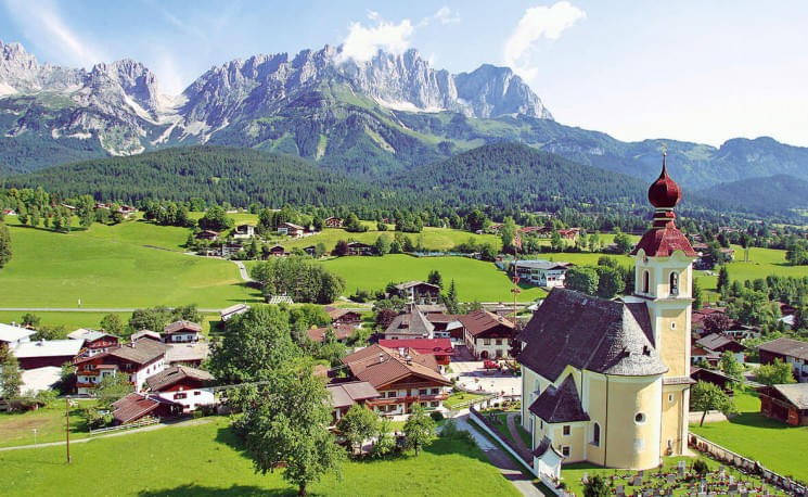 Ein malerisches Bergdorf mit traditionellen Häusern, einer Kirche und einem Bergpanorama im Hintergrund. Grüne Wiesen und ein klarer Himmel runden das Bild ab.