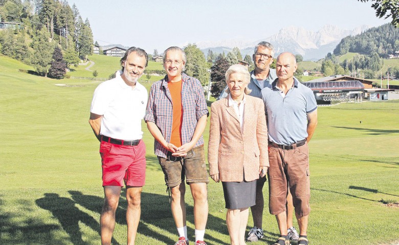 Eine Gruppe von fünf Menschen steht auf einem sonnigen Golfplatz mit Bergen im Hintergrund. Alle lächeln in lässiger Kleidung in die Kamera.