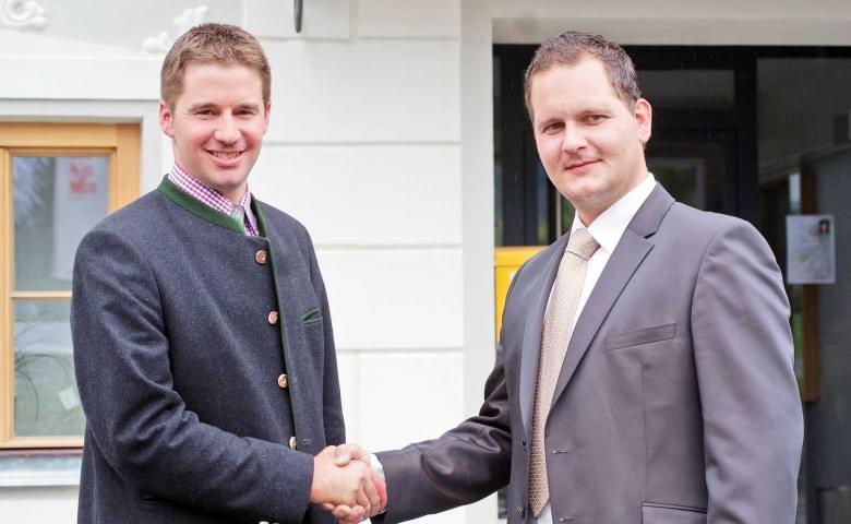 Zwei Männer in formeller Kleidung schütteln sich vor einem Gebäude die Hände, beide lächeln freundlich. Einer trägt einen Anzug, der andere eine traditionelle Jacke.