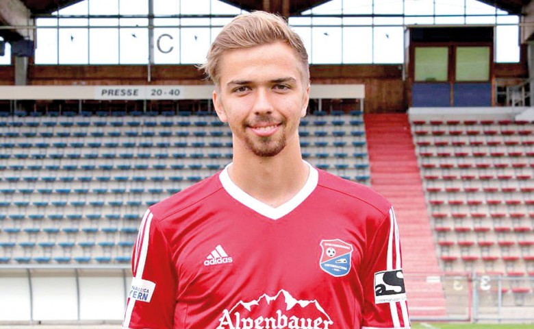 Ein Fußballspieler in einem roten Trikot steht auf einem Spielfeld vor leeren Rängen. Er lächelt und trägt ein Trikot mit der Aufschrift "Alpenbauer".