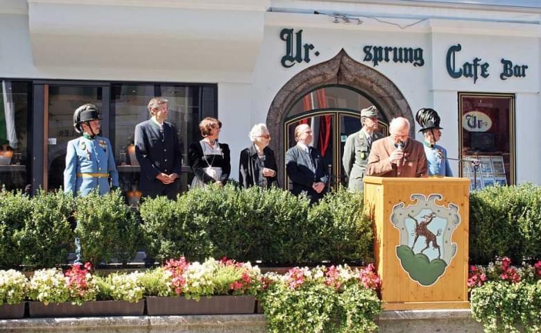 Eine Gruppe von Menschen steht auf einer Bühne vor einem Café. Ein Mann hält eine Rede, während andere formell gekleidet zuhören. Blumen dekorieren die Bühne.