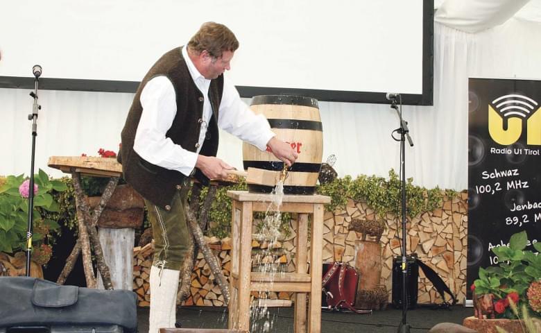 Ein Mann in traditioneller Kleidung zapft ein Fass Bier auf einer Bühne mit Mikrofonen und dekorativem Hintergrund aus Holz und Pflanzen.