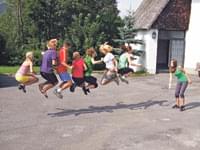 Gruppenspringen: Mehrere Kinder und Jugendliche springen synchron auf einem Asphaltplatz vor einer Hütte in die Höhe, alle lächeln und haben Spaß.