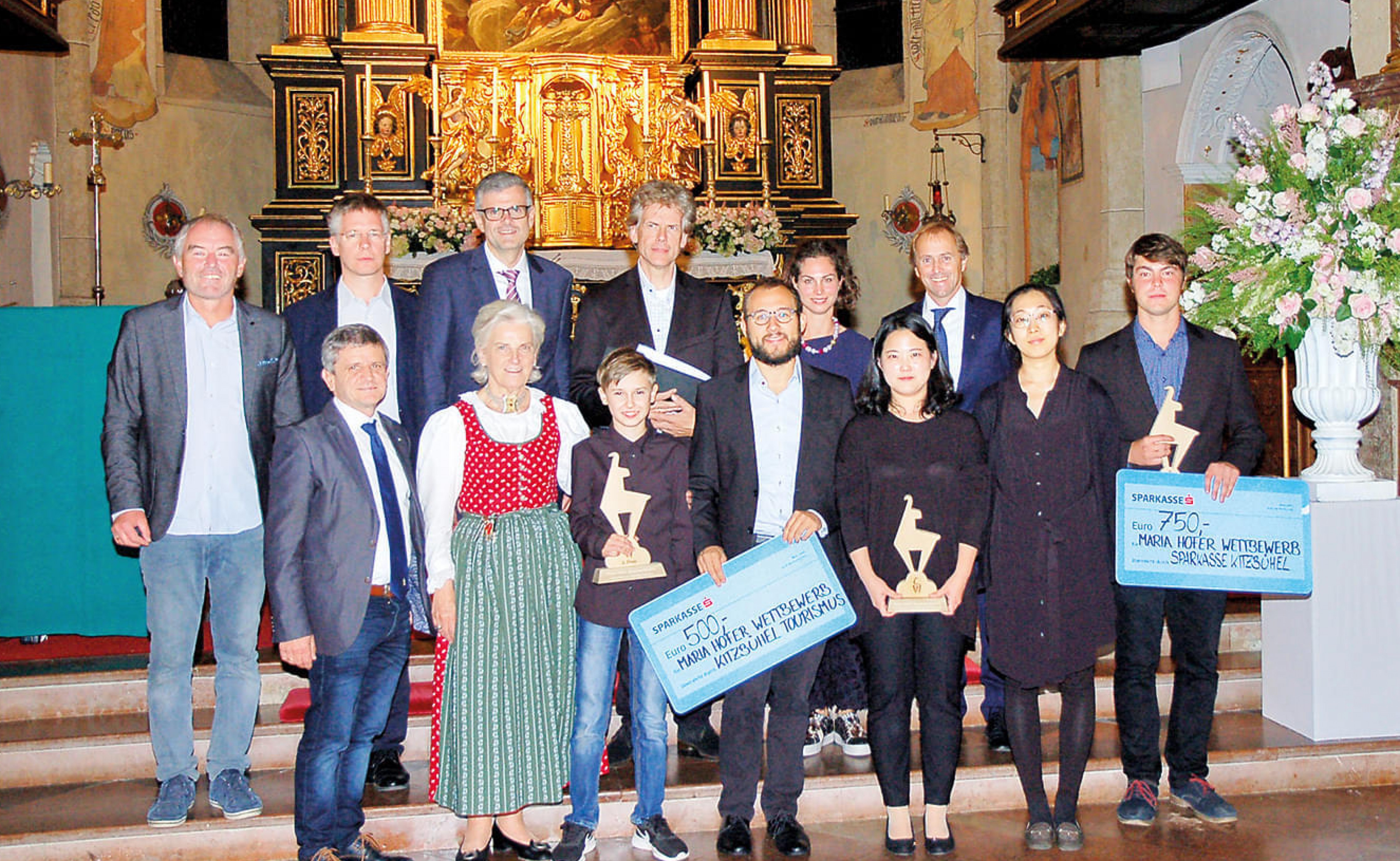 Gruppe von Menschen in festlicher Kleidung posiert in einer Kirche mit Dekorationen und Trophäen. Zwei Personen halten blaue Schilder mit Text.