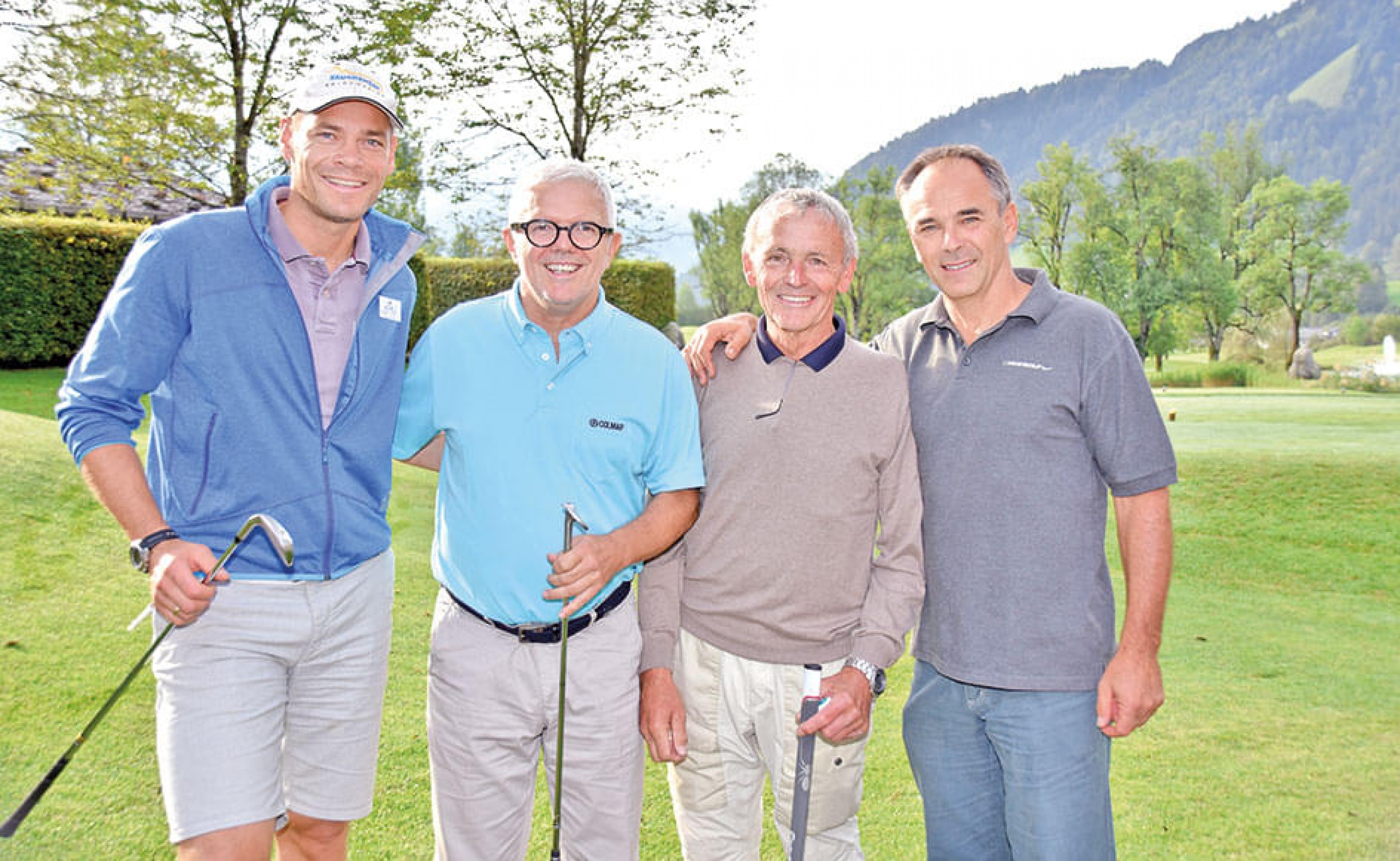 Vier Männer stehen lächelnd auf einem Golfplatz, jeder hält einen Golfschläger. Sie tragen sportliche Freizeitkleidung und genießen das sonnige Wetter.