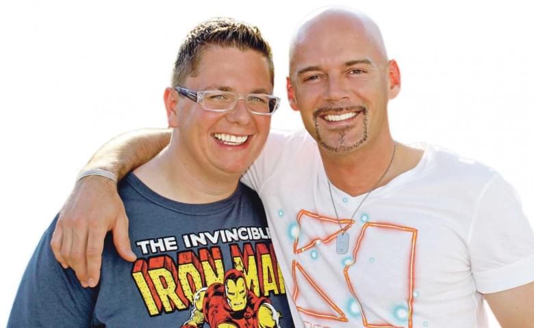 Zwei Männer lächeln in die Kamera. Der linke trägt ein Iron Man T-Shirt, der rechte ein weißes Shirt. Sie haben die Arme umeinander gelegt.