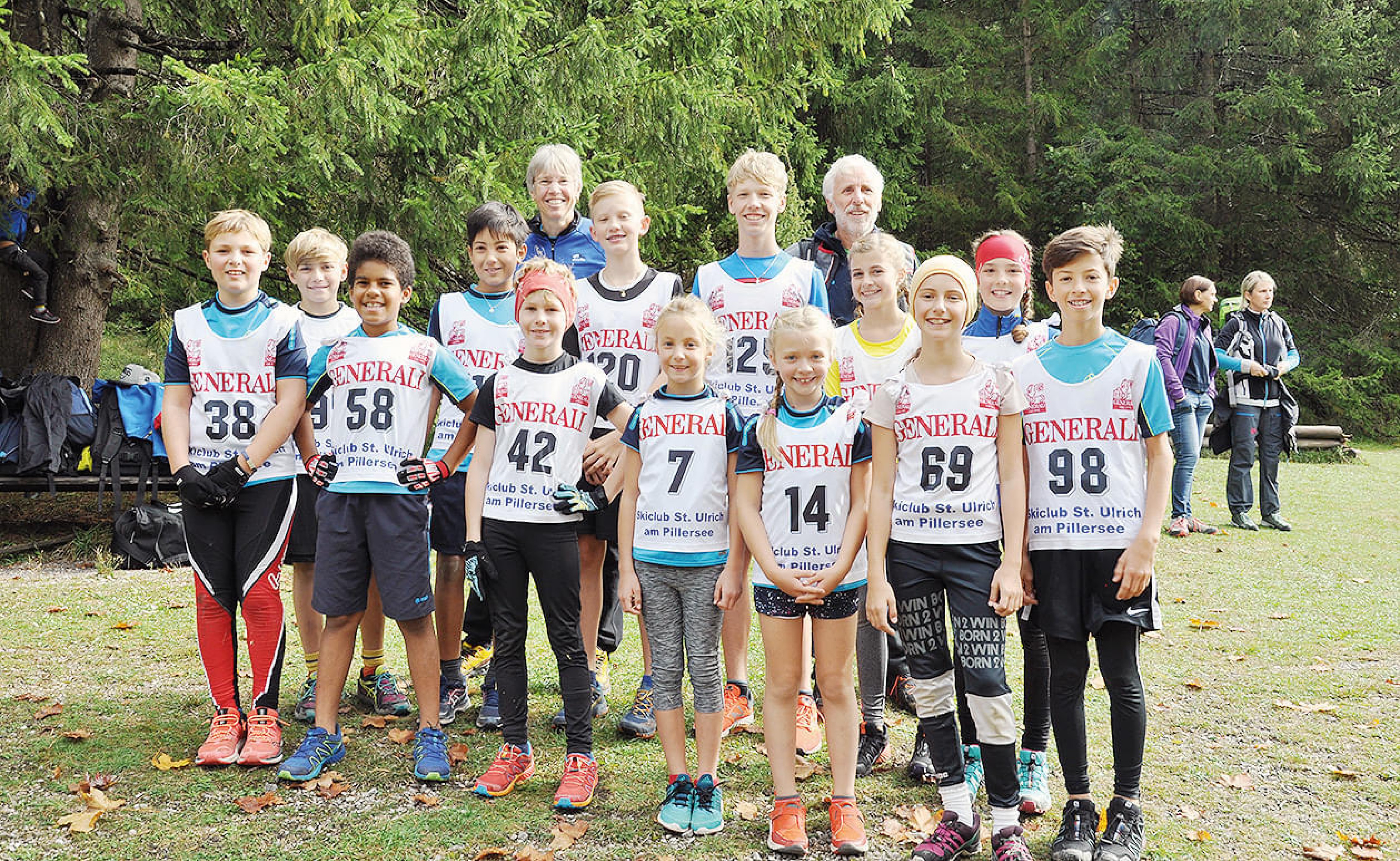Eine Gruppe von jungen Läufern in nummerierten Trikots posiert lächelnd draußen auf einer Wiese. Erwachsene stehen dahinter, Bäume im Hintergrund.