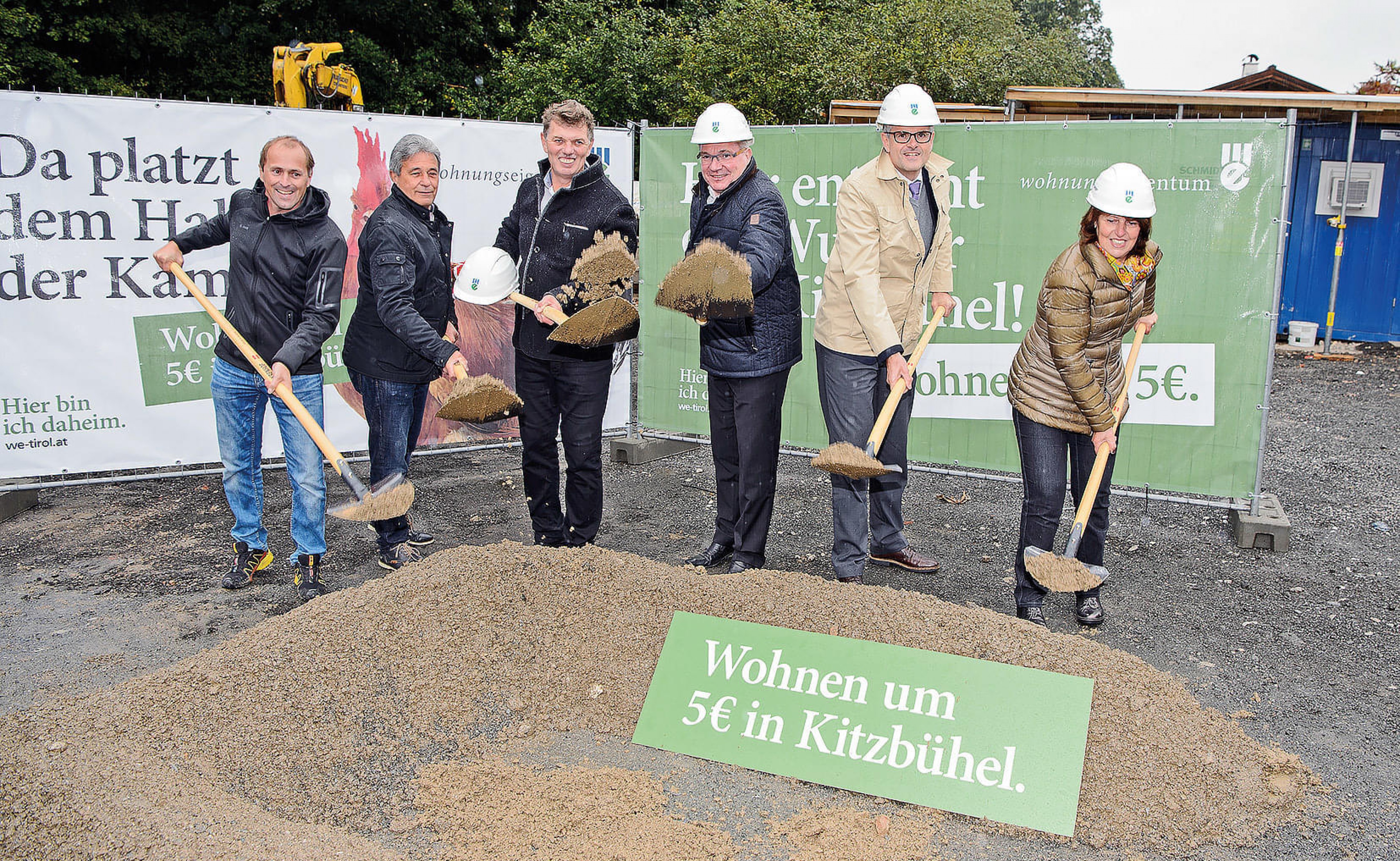 Gruppe von Personen mit Bauhelmen hält Schaufeln bei einer Grundsteinlegung; Schilder werben für günstiges Wohnen ab 5 Euro in Kitzbühel.