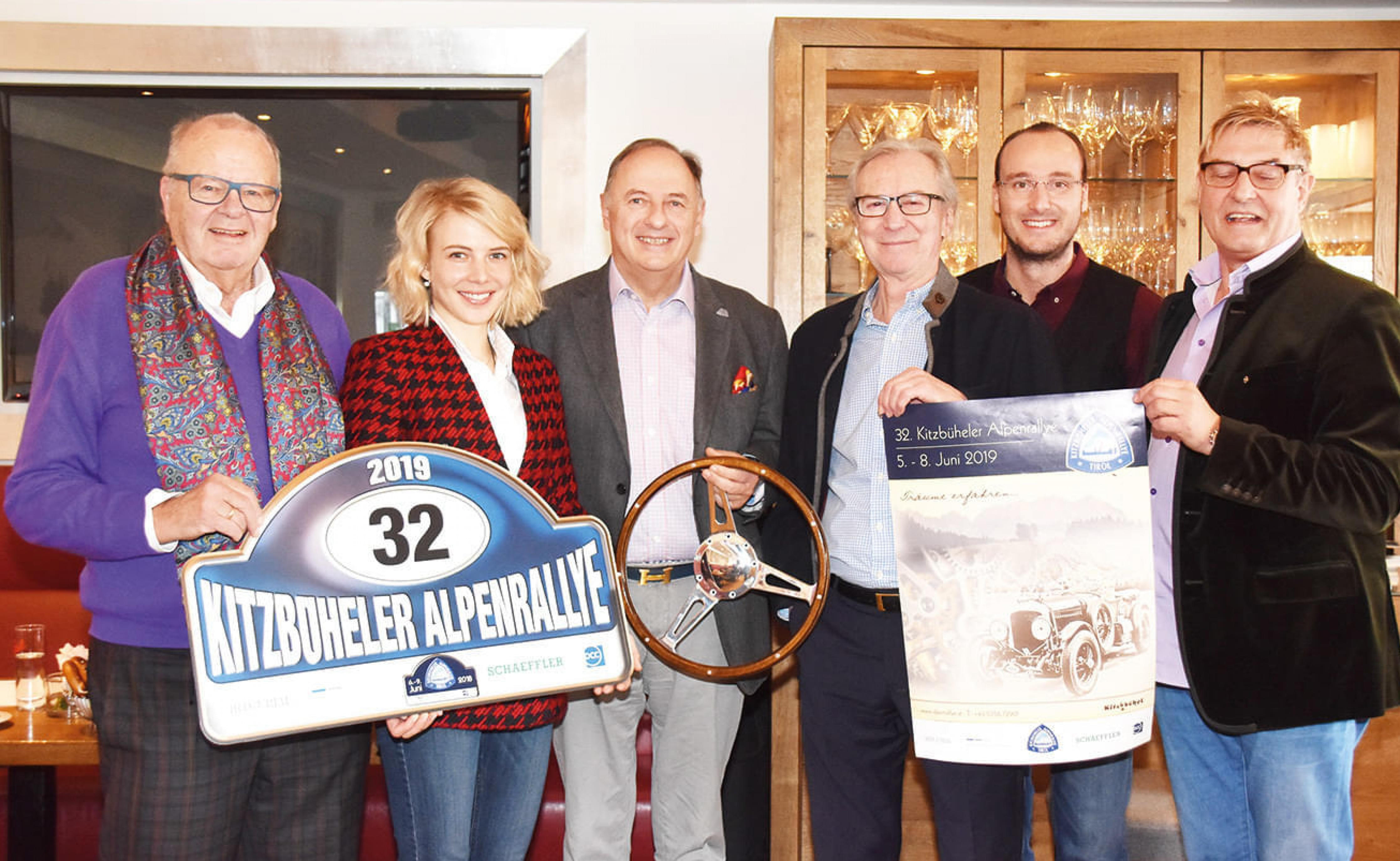 Sechs Personen halten ein großes Schild mit der Aufschrift 'Kitzbüheler Alpenrallye', sowie ein Lenkrad und ein Plakat mit einem Oldtimer-Motiv.