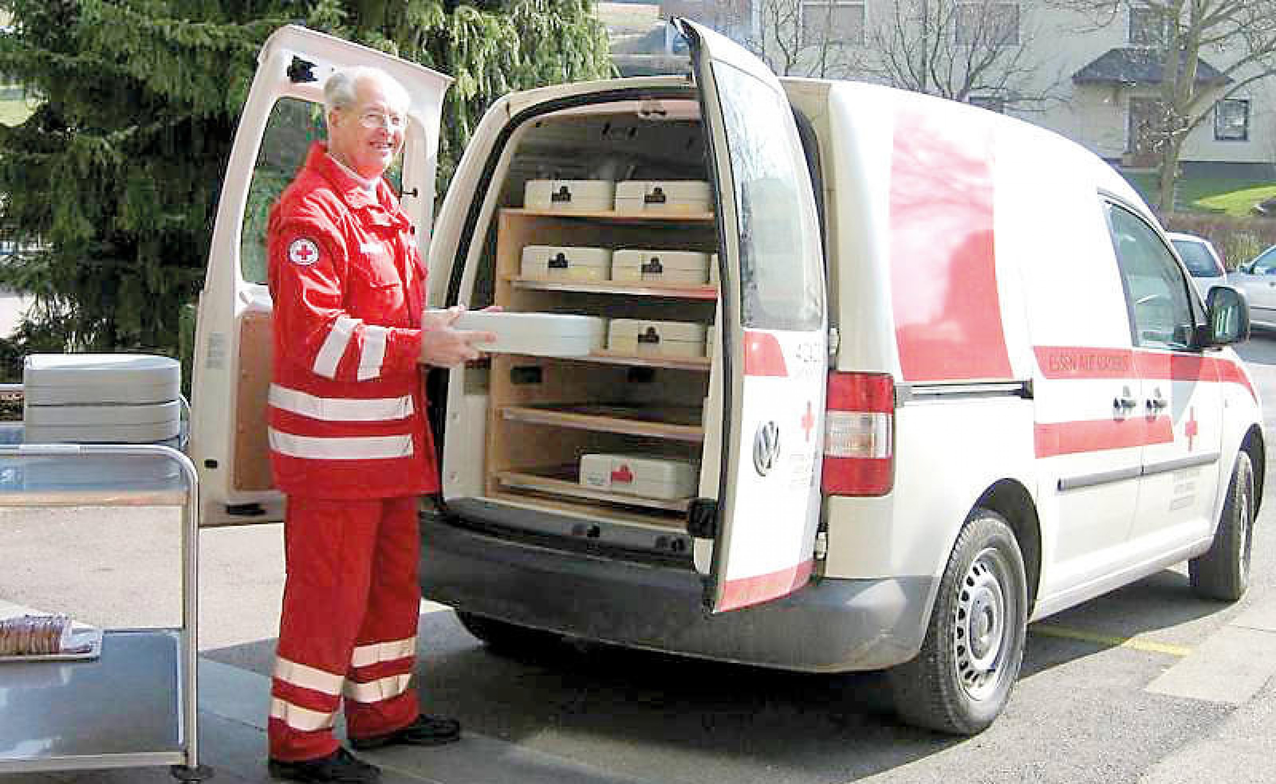 Ein Mann in roter Arbeitskleidung steht neben einem Lieferwagen mit geöffneter Hecktür. Der Wagen enthält mehrere Kisten, vermutlich für Versorgungsgüter.