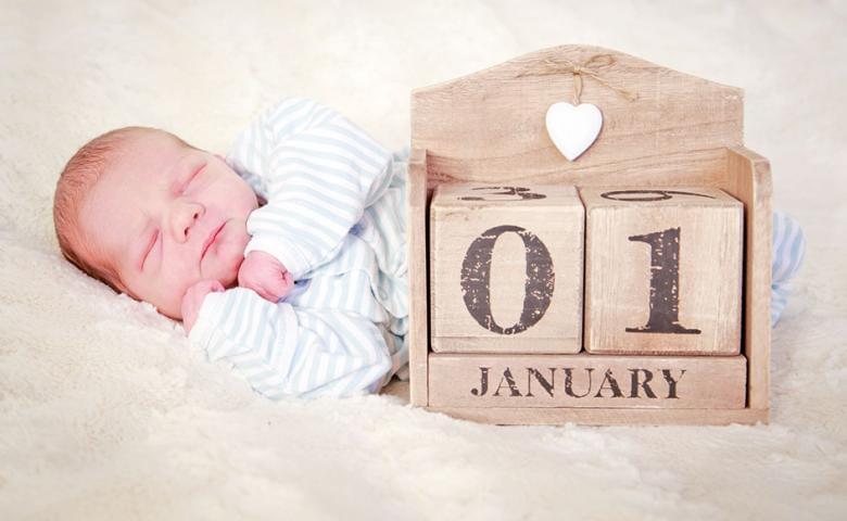 Ein schlafendes Baby in Streifenpyjama liegt neben Holzblöcken, die das Datum "01 Januar" zeigen. Der Hintergrund ist weich und flauschig.