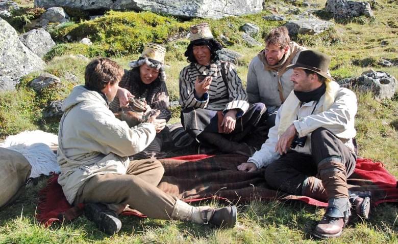 Eine Gruppe von fünf Personen sitzt in historischer Kleidung auf einer Wiese und isst. Sie sind von Felsen und Gras umgeben, während sie in einer Runde plaudern.