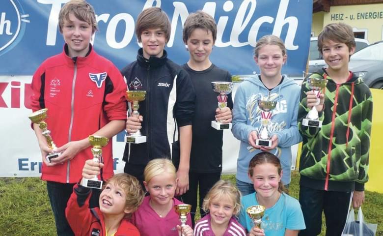 Gruppe junger Menschen, die Pokale vor einem Banner mit der Aufschrift "Tirol Milch" halten, lächeln in die Kamera. Sie tragen Freizeitkleidung.