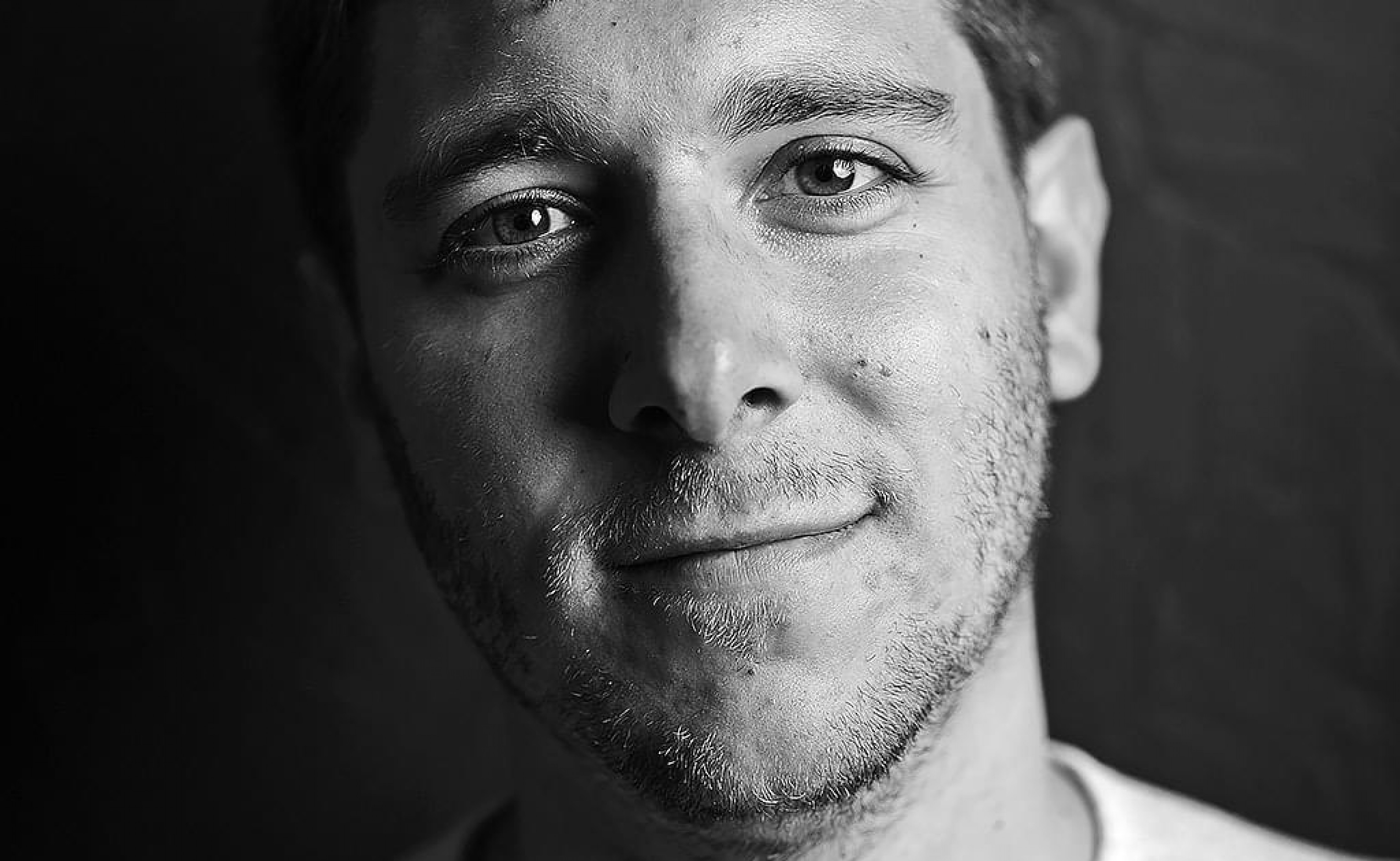 Porträt eines Mannes mit kurzem Haar und Bart, der leicht lächelt und direkt in die Kamera schaut, in Schwarzweiß gehalten.