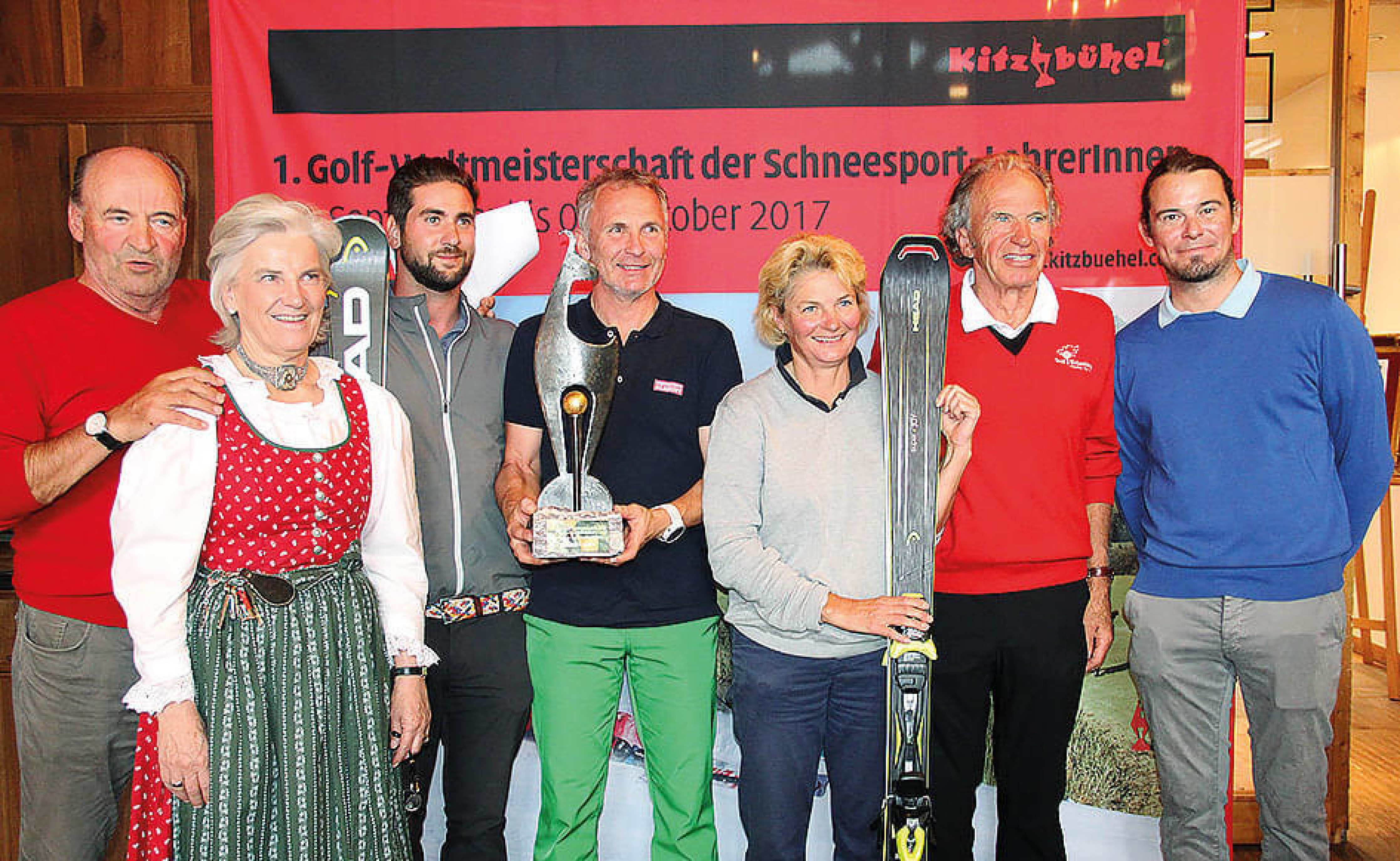 Eine Gruppe von sieben Personen posiert mit Skiern und einem Pokal vor einem Banner für ein Schneesport-Golf-Event in Kitzbühel im Jahr 2017.