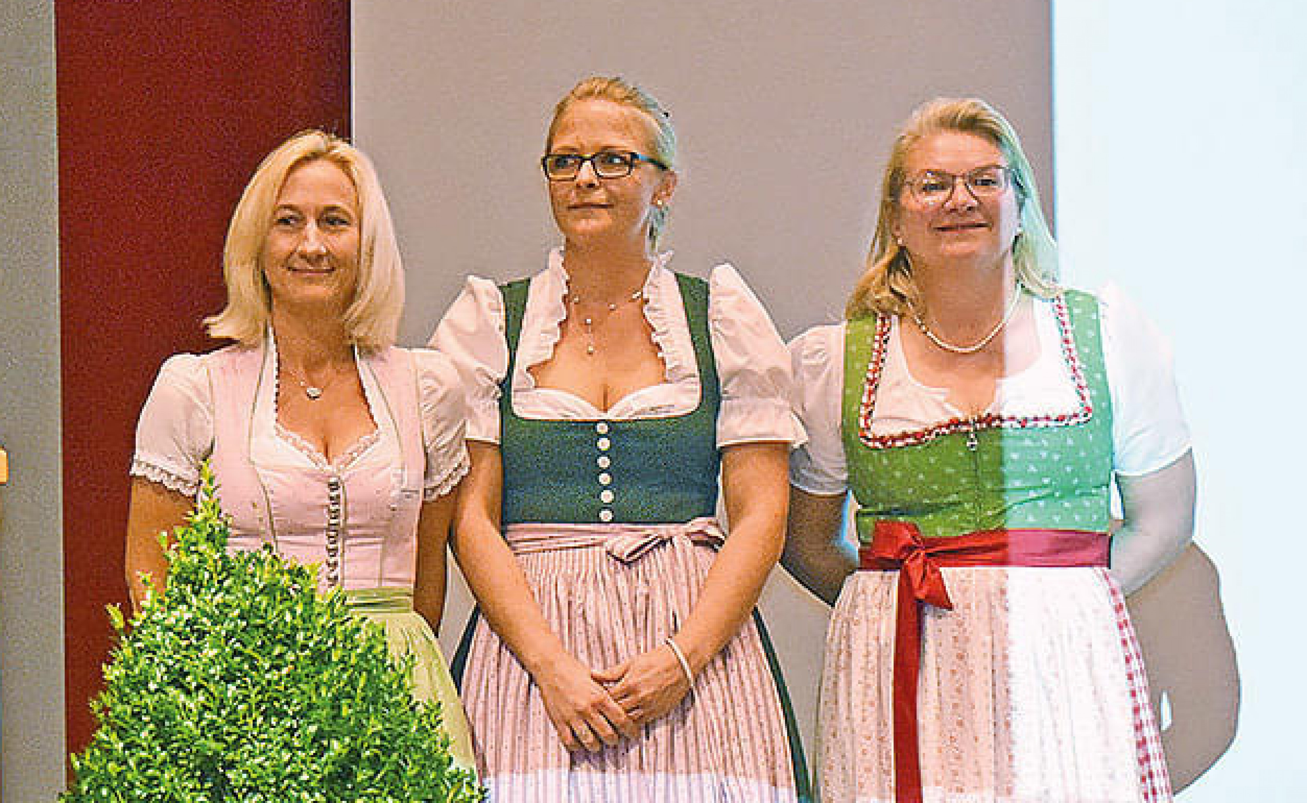 Drei Frauen in traditionellen bayerischen Dirndln stehen nebeneinander vor einem roten und weißen Hintergrund. Eine grüne Pflanze ist im Vordergrund.