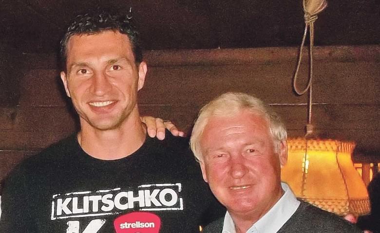 Zwei Männer stehen lächelnd nebeneinander in einem Raum mit Holzverkleidung und einer Hängelampe.