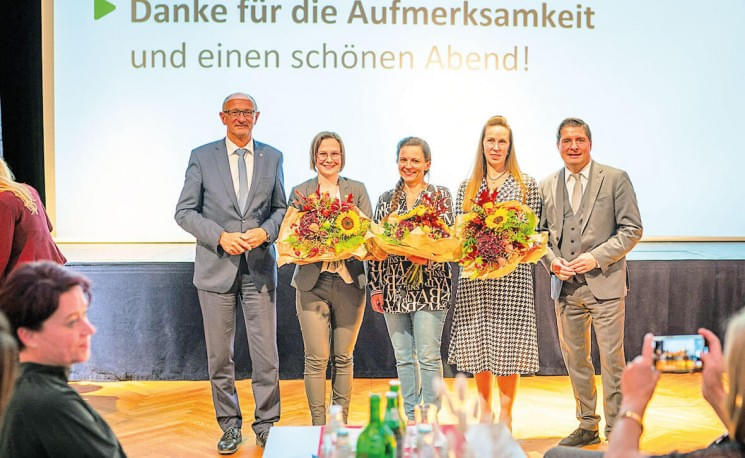 Fünf Personen stehen auf einer Bühne, drei Frauen mit Blumensträußen in der Mitte. Im Hintergrund ist ein Dankesschriftzug zu sehen.