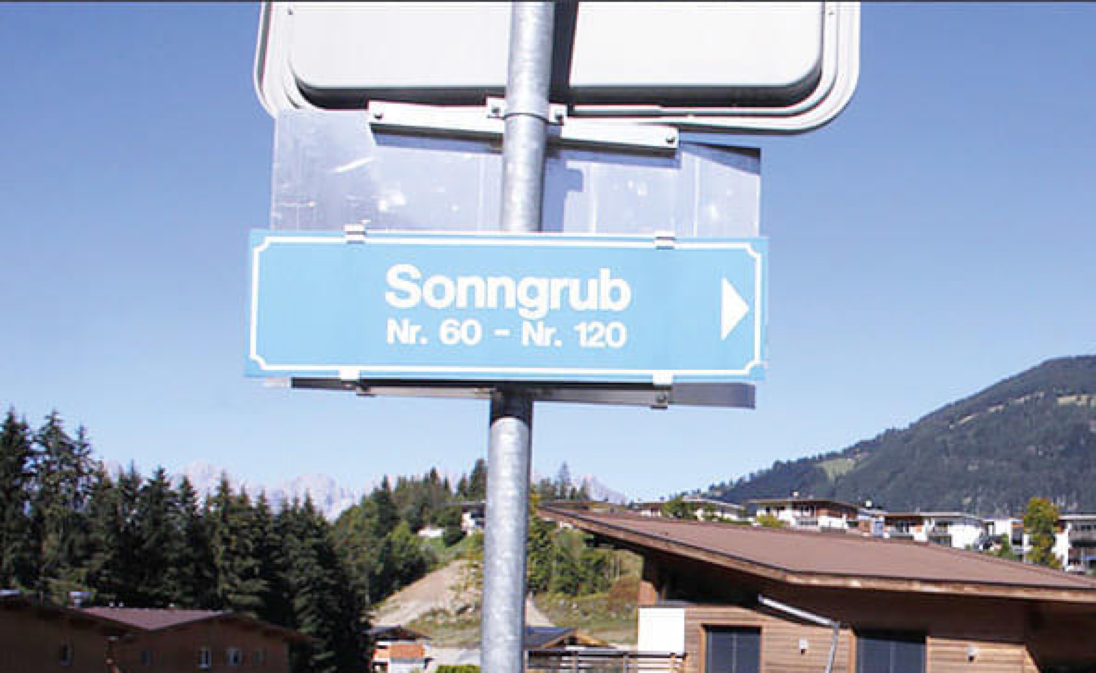 Ein Straßenschild mit der Aufschrift "Sonngrub Nr. 60 - Nr. 120" zeigt nach rechts, umgeben von Bergen und Bäumen unter einem klaren blauen Himmel.