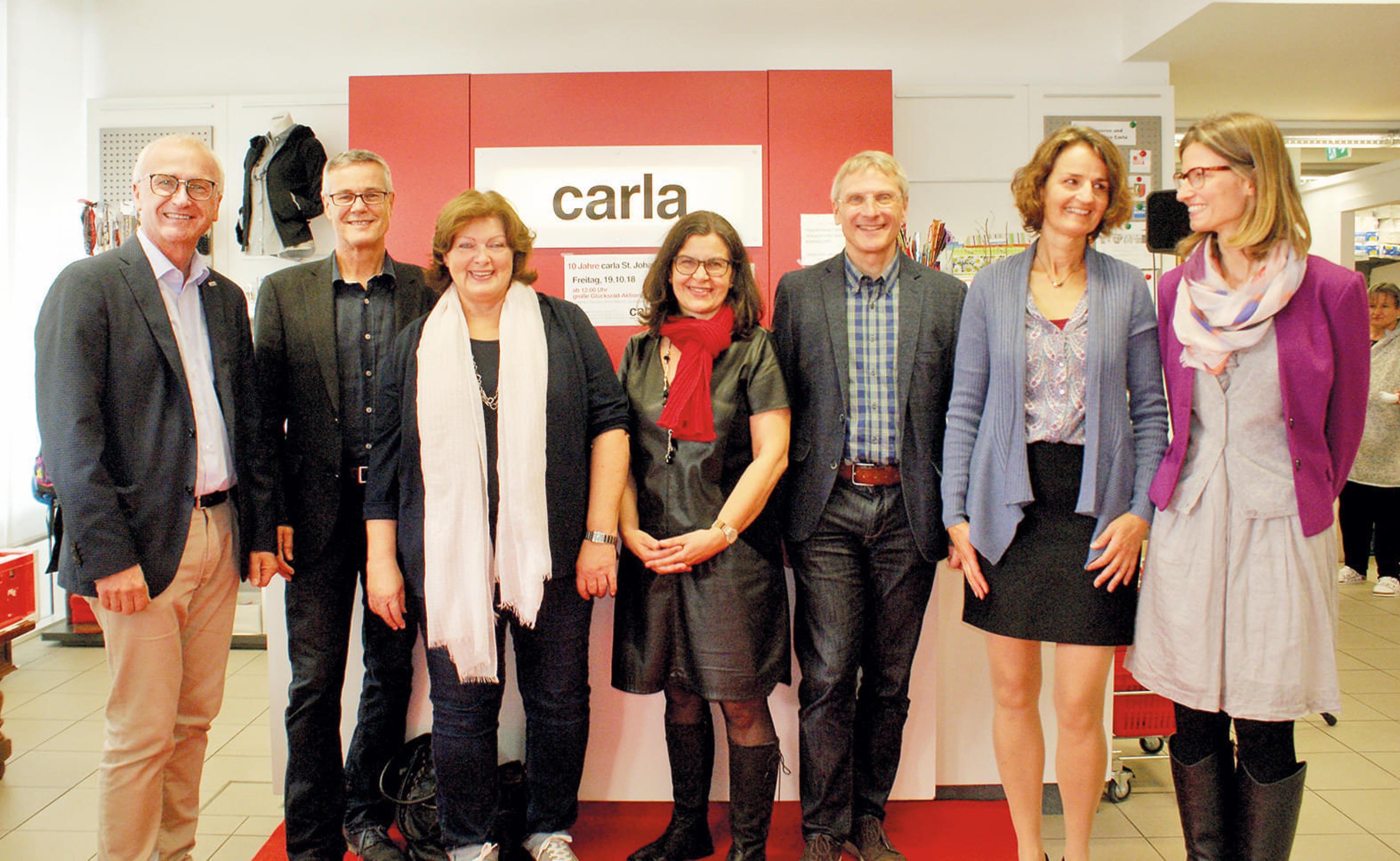 Eine Gruppe von sieben Personen steht lächelnd nebeneinander vor einer roten Wand mit einem Schild "carla". Alle sind lässig bis elegant gekleidet.