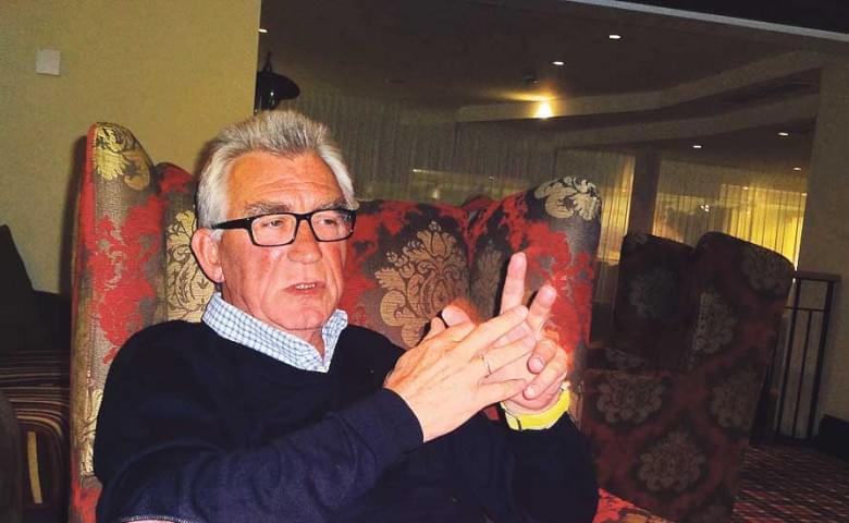 Älterer Mann mit Brille und grauen Haaren sitzt in einem gemusterten Sessel und gestikuliert mit den Händen. Im Hintergrund sind Vorhänge sichtbar.