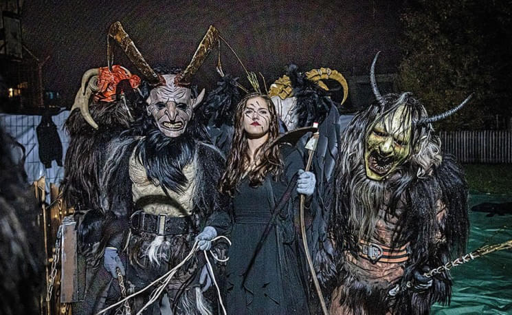 Person mit zwei dämonischen Gestalten auf einem nächtlichen Kostümfest, beide tragen furchterregende Masken und Hörner.