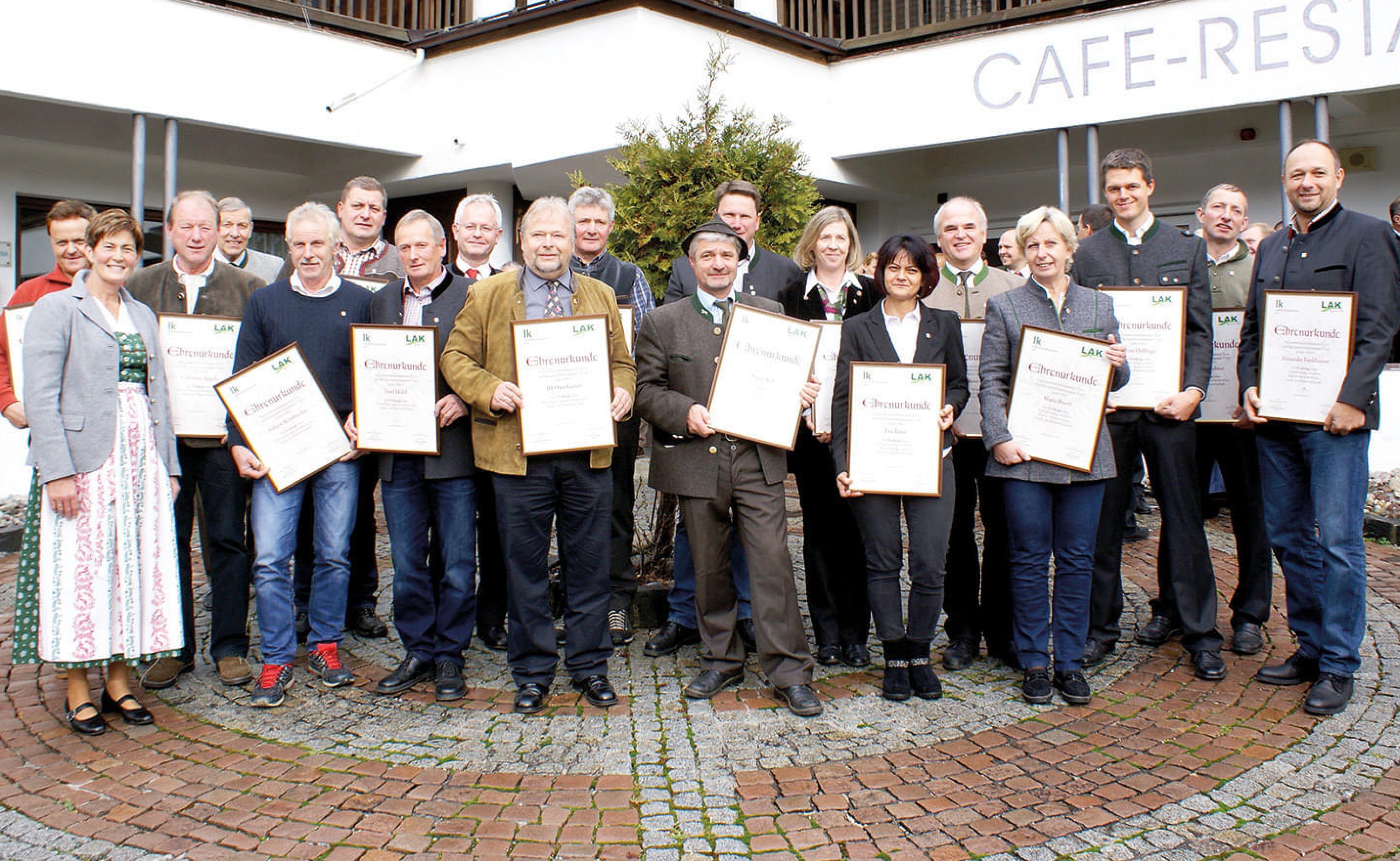 Eine Gruppe von Menschen steht vor einem Gebäude und hält Urkunden in den Händen, lächelnd in die Kamera. Es scheint eine feierliche Veranstaltung zu sein.