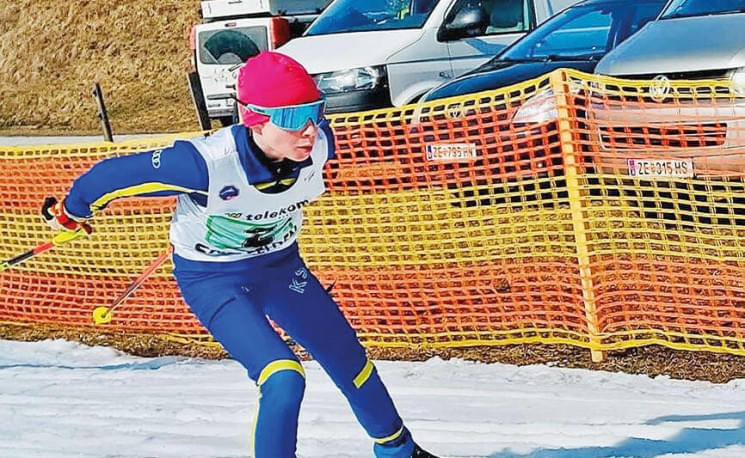 Langläuferin in blauer und gelber Ausrüstung, roter Mütze und Sonnenbrille, fährt auf Schnee. Orange Absperrung und Autos im Hintergrund.