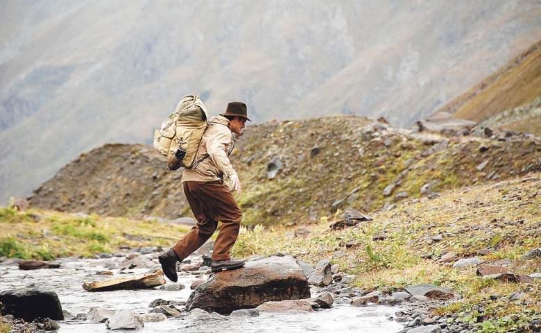 Eine Person in Wanderkleidung und Hut überquert einen kleinen Fluss auf Steinen in einer bergigen Landschaft, umgeben von kahlen, felsigen Hügeln.