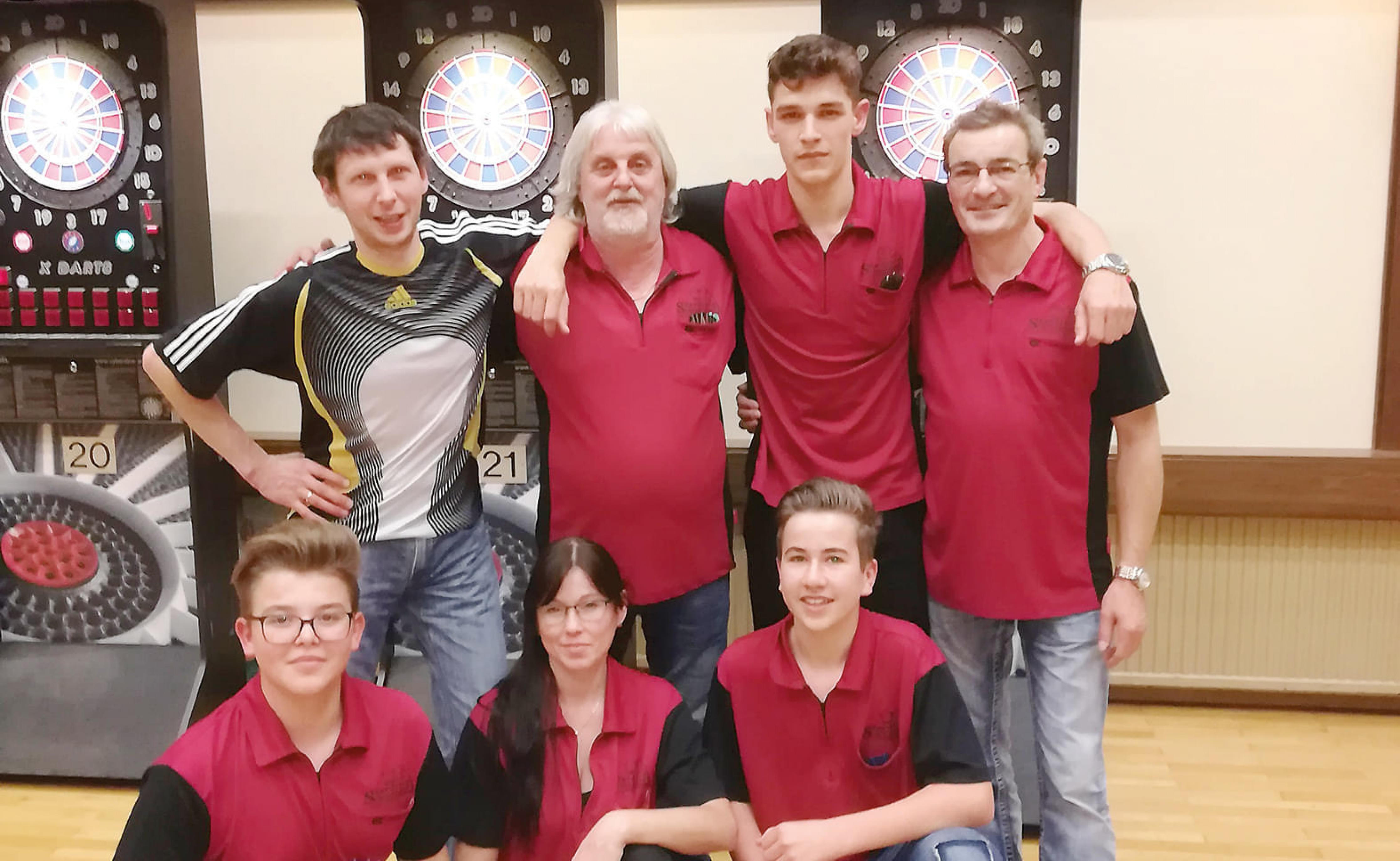 Sieben Personen posieren lächelnd vor elektronischen Dartscheiben in einem Raum. Einige tragen rote Hemden, andere Freizeitkleidung. Die Stimmung wirkt entspannt.