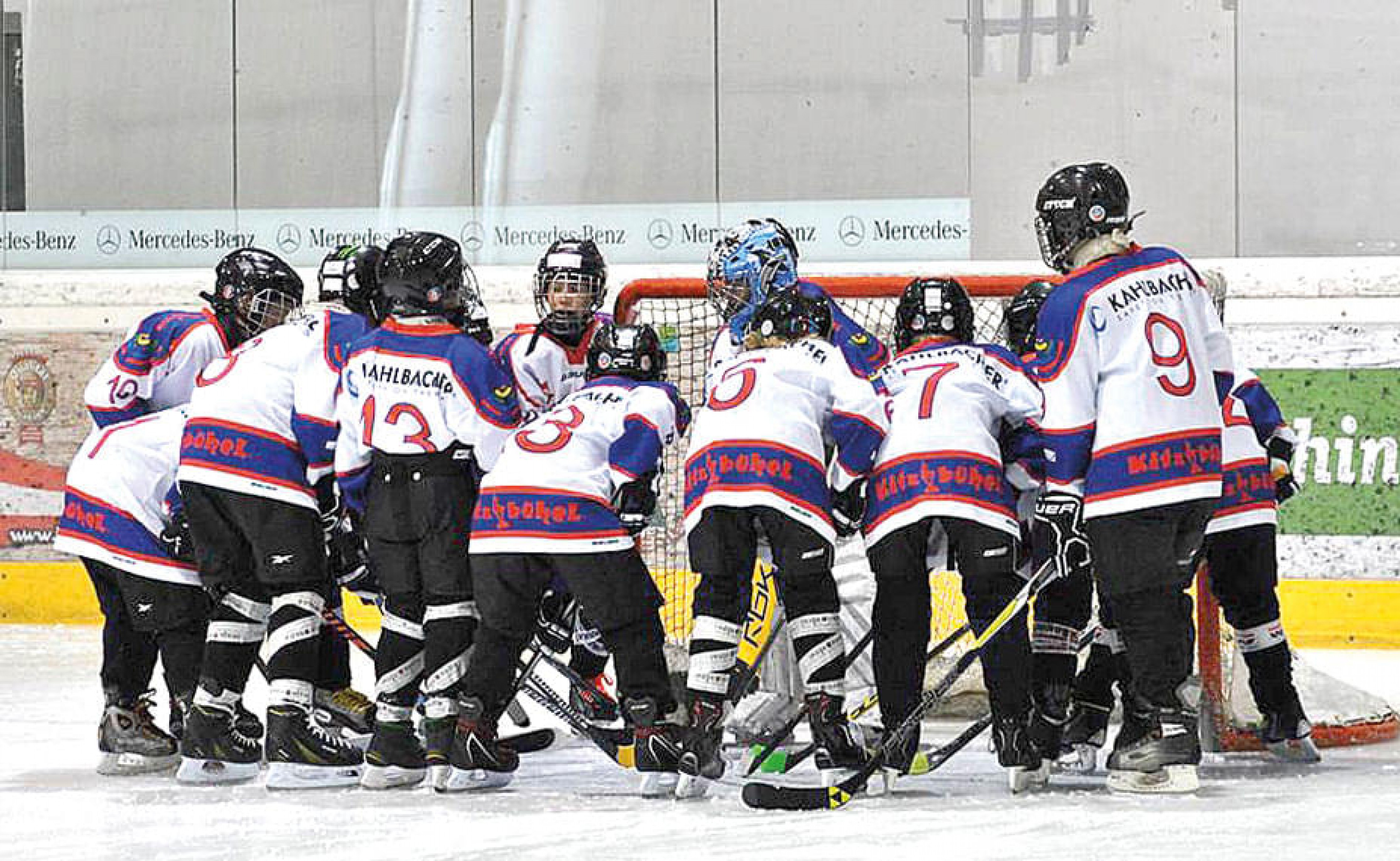 Eine Gruppe von jungen Eishockeyspielern in weißen Trikots versammelt sich eng um ein Tor auf dem Eis eines Eishockeyfeldes.