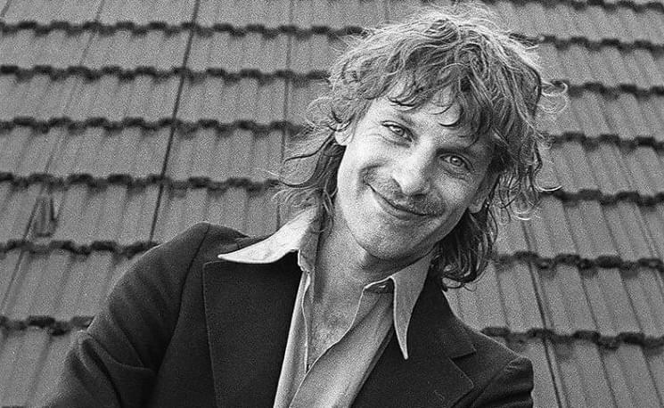 Schwarz-weiß Porträt eines lächelnden Mannes mit längeren Haaren und einem Anzug vor einer Ziegelwand.