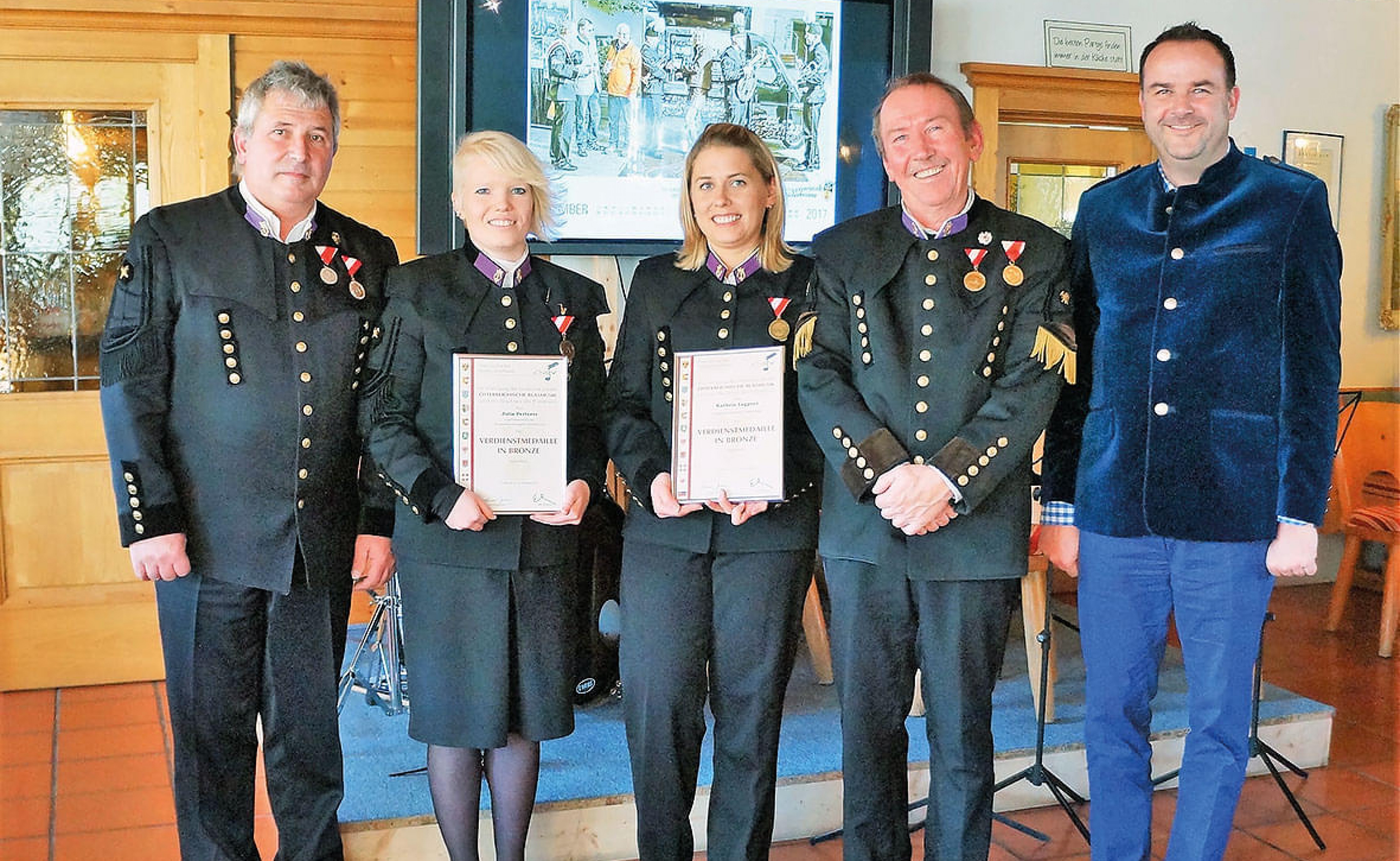Fünf Personen in festlicher Kleidung stehen lächelnd in einem Raum, zwei von ihnen halten Urkunden, und alle tragen Medaillen an ihren Jacken.