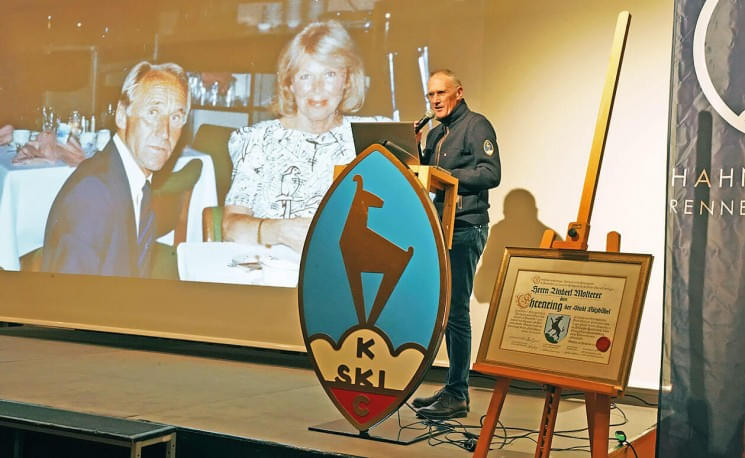 Ein Mann steht auf einer Bühne neben einem großen Ski-Logo und einer gerahmten Urkunde. Im Hintergrund ist ein Foto von einer älteren Paar zu sehen.