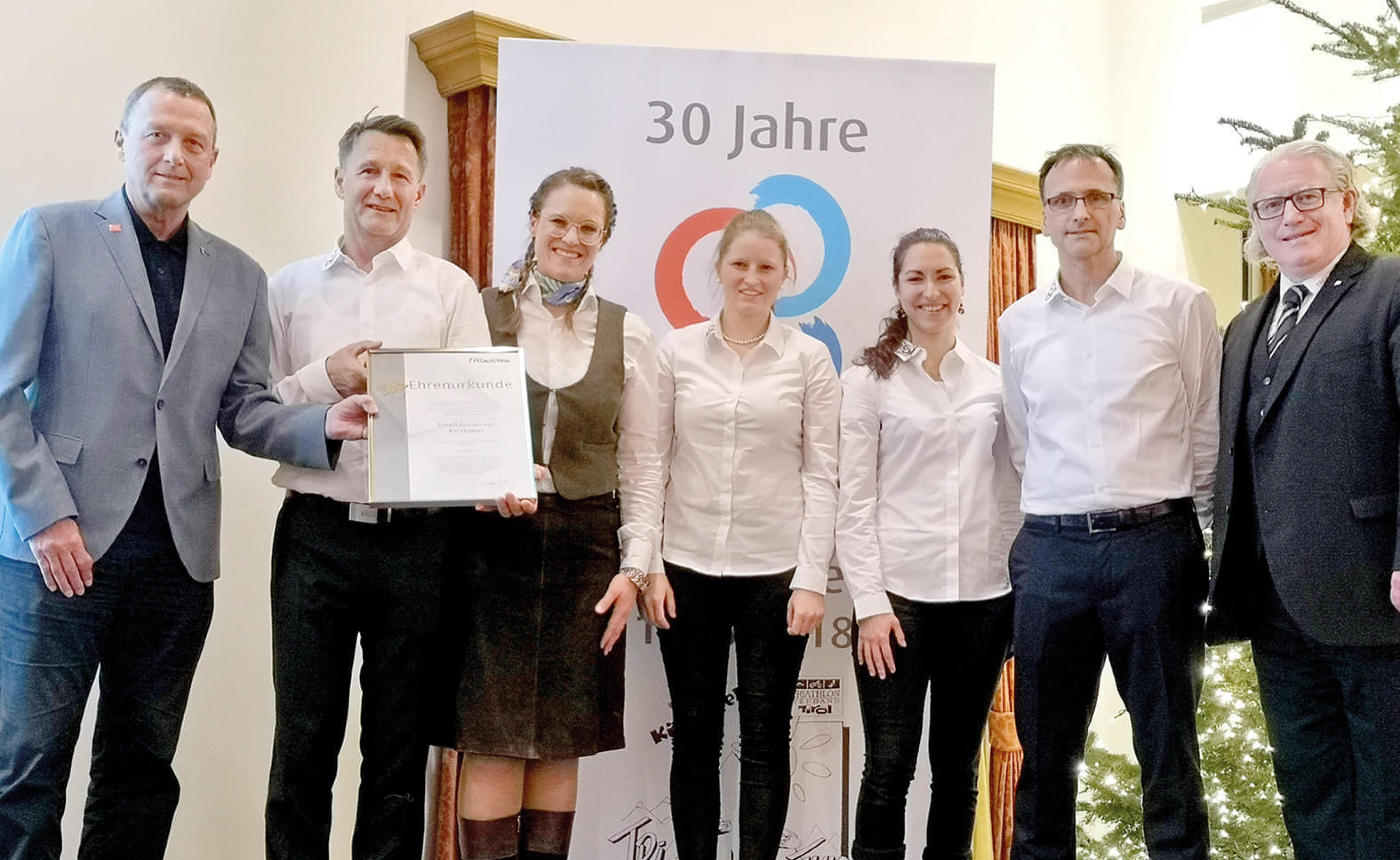 Eine Gruppe von sieben Personen steht vor einem Banner mit der Aufschrift "30 Jahre" und lächelt in die Kamera. Zwei Personen halten ein Zertifikat.