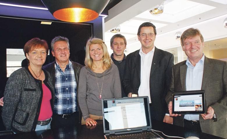 Sechs Menschen stehen lächelnd hinter einem Tisch mit einem offenen Laptop und einem Tablet. Sie tragen legere und businessmäßige Kleidung.