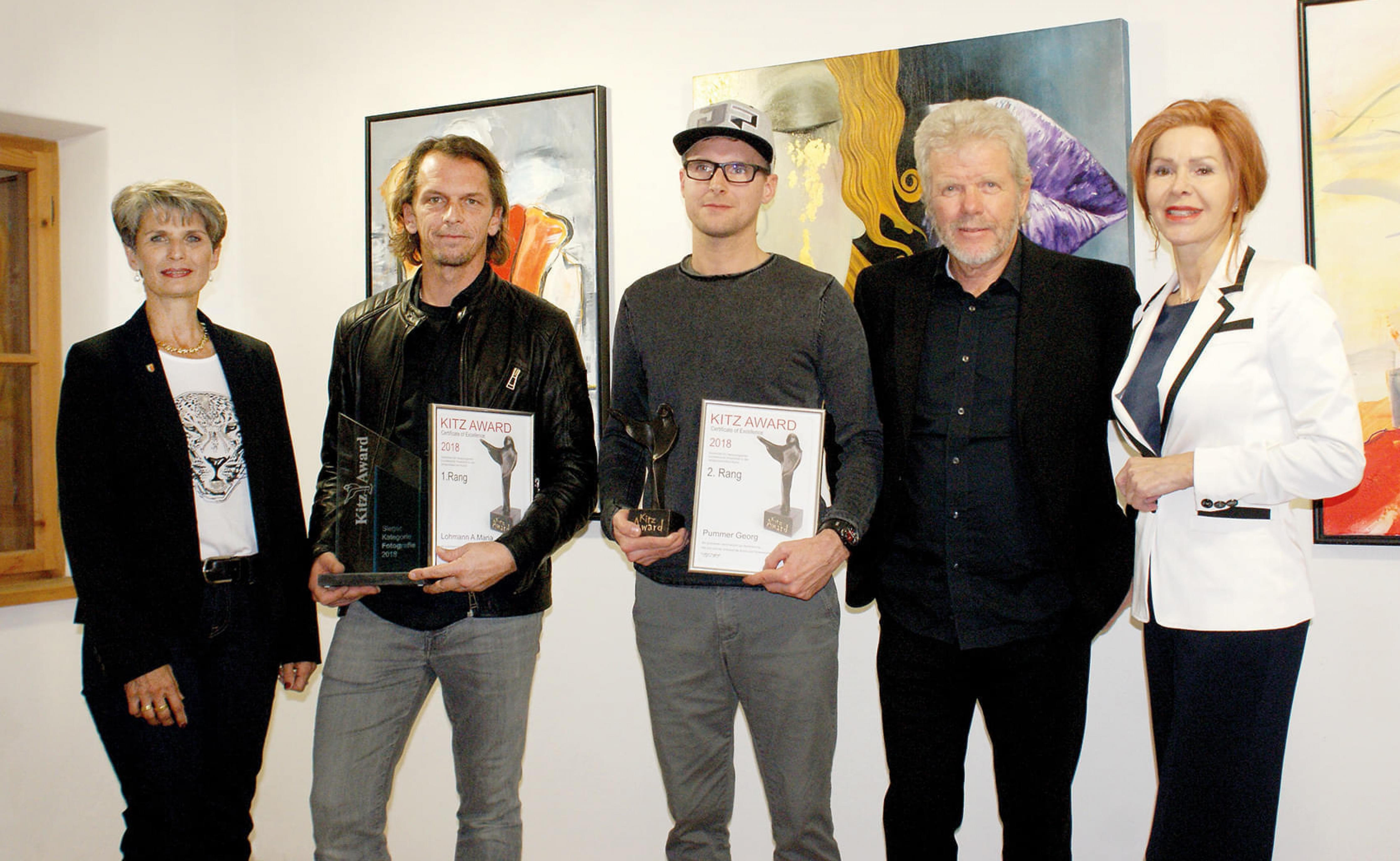 Fünf Personen posieren vor Kunstwerken an einer Wand, wobei zwei von ihnen Auszeichnungen halten. Alle tragen semi-formelle Kleidung und lächeln in die Kamera.