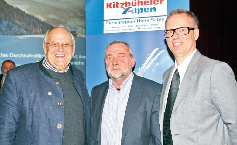 Drei Männer in Anzügen stehen nebeneinander und lächeln in die Kamera. Im Hintergrund sind Banner mit Texten und Logos sichtbar.