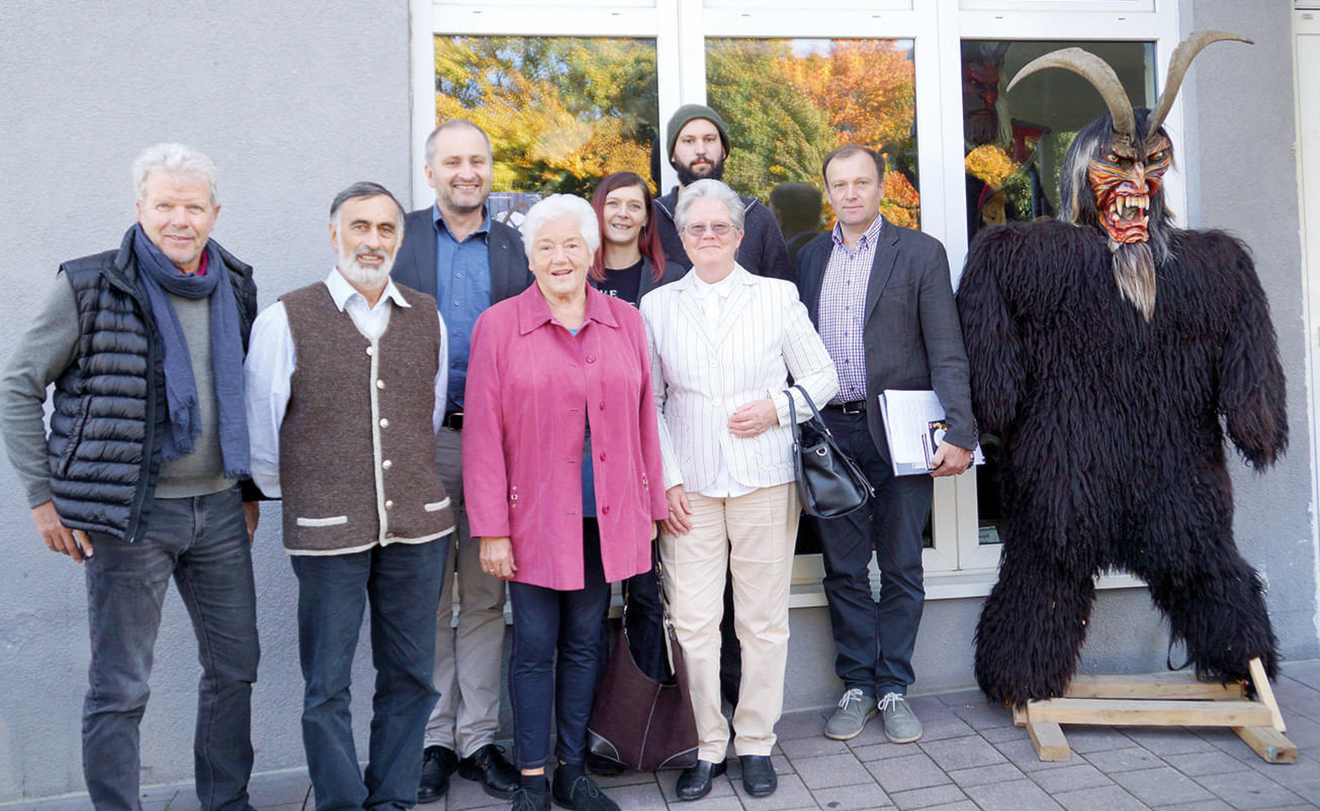 Gruppe von Menschen steht vor einem Gebäude, eine Person trägt ein Krampus-Kostüm. Herbstliche Bäume spiegeln sich im Fenster wider. Alle lächeln in die Kamera.