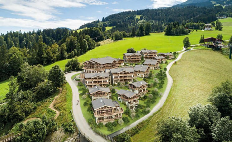 Luftaufnahme einer malerischen Ferienanlage inmitten grüner Hügel und Wälder. Traditionelle Holzhäuser gruppieren sich entlang geschwungener Straßen.