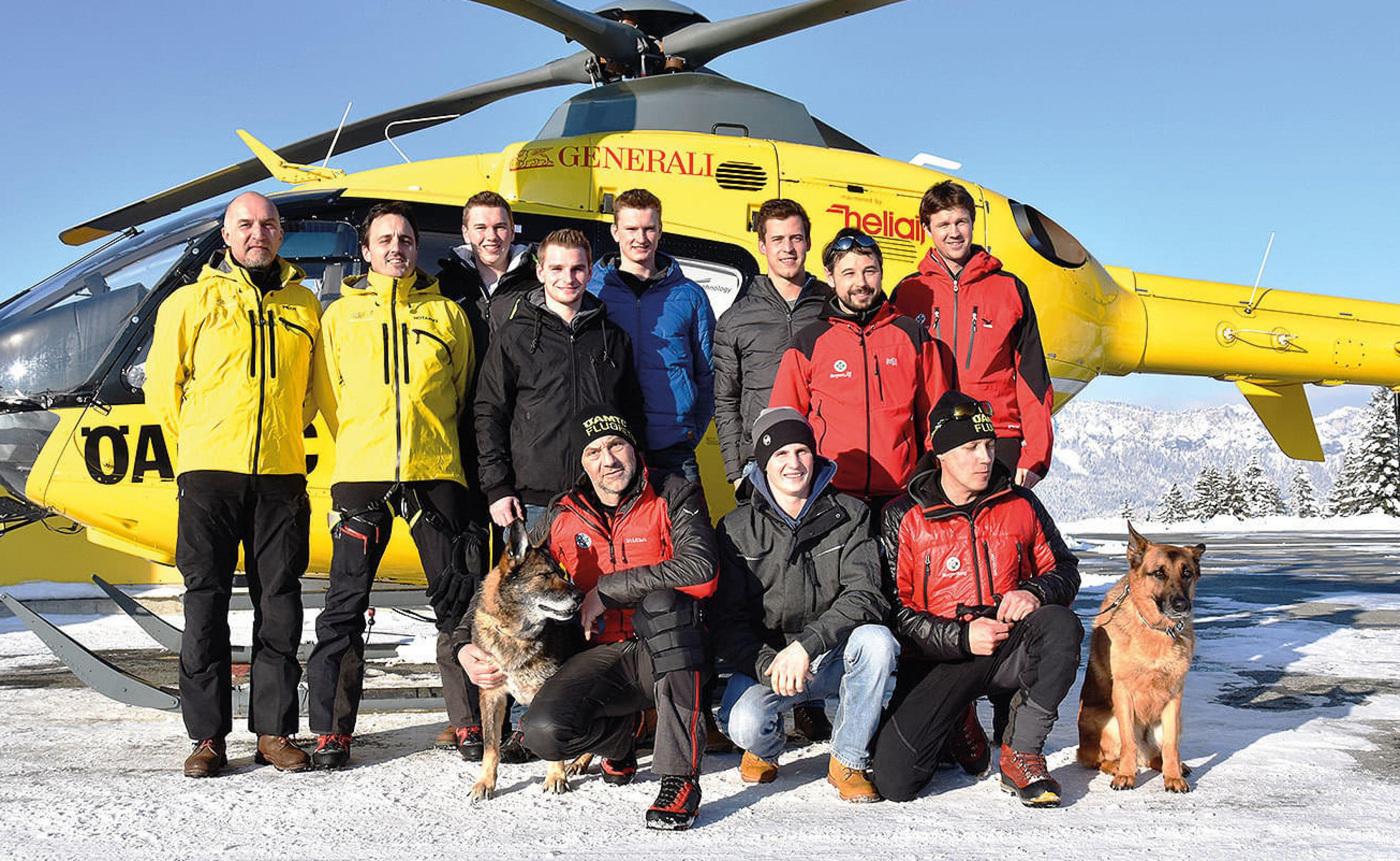Gruppe von Männern in Winterkleidung posiert lächelnd vor einem gelben Rettungshubschrauber in schneebedeckter Landschaft, begleitet von zwei Schäferhunden.