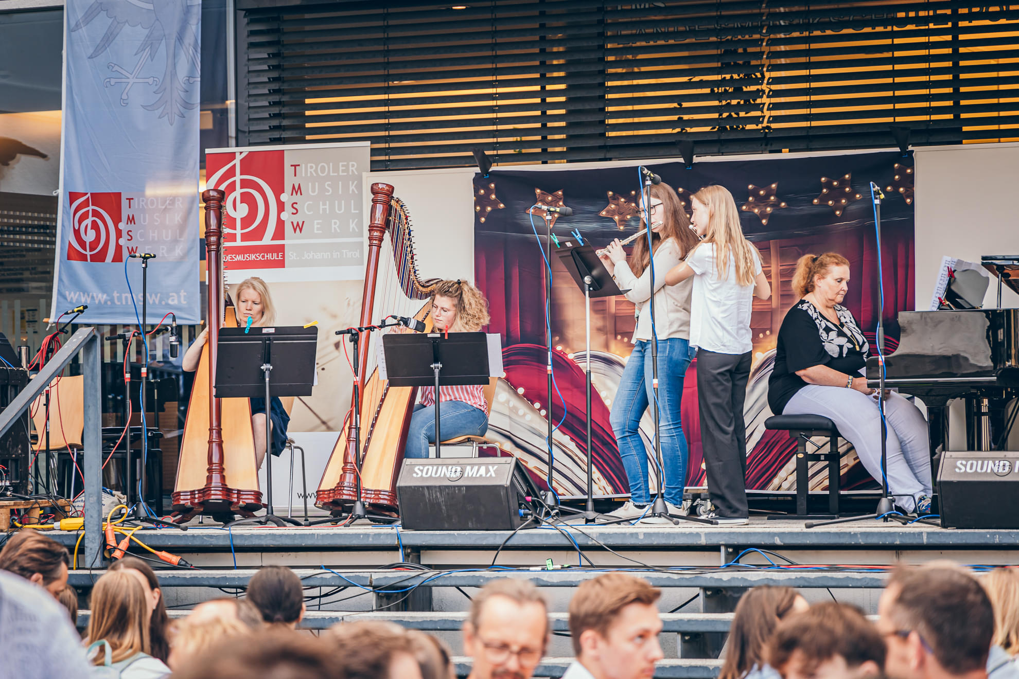 Eine Gruppe junger Musikerinnen und Musiker spielt ein Konzert auf einer Freilichtbühne, umgeben von Publikum. Es sind Harfen und ein Klavier zu sehen.