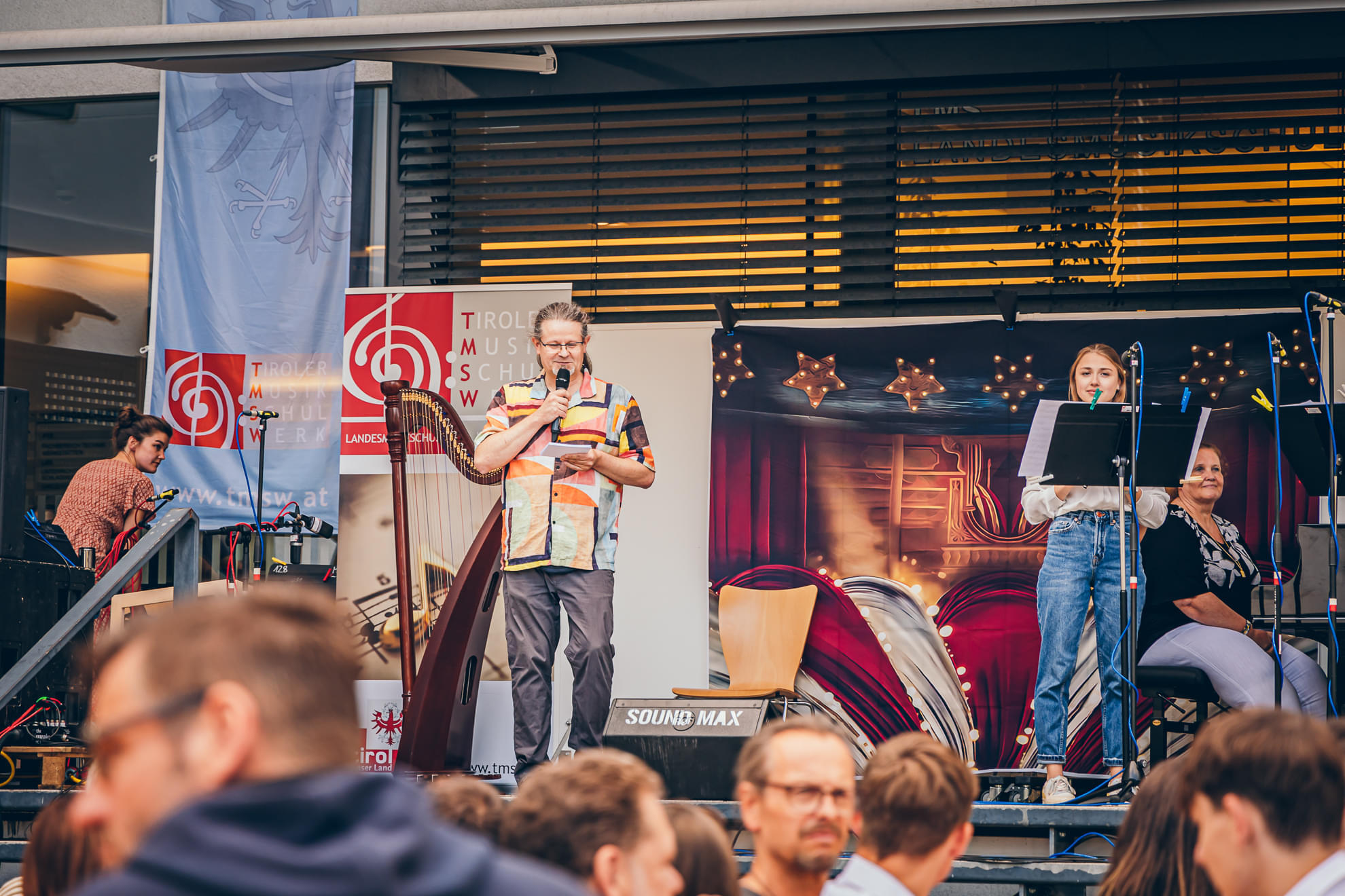 Eine Live-Bühnenperformance mit einem Moderator und einer Sängerin, umgeben von Musik-Equipment. Zuschauer schauen interessiert zu.