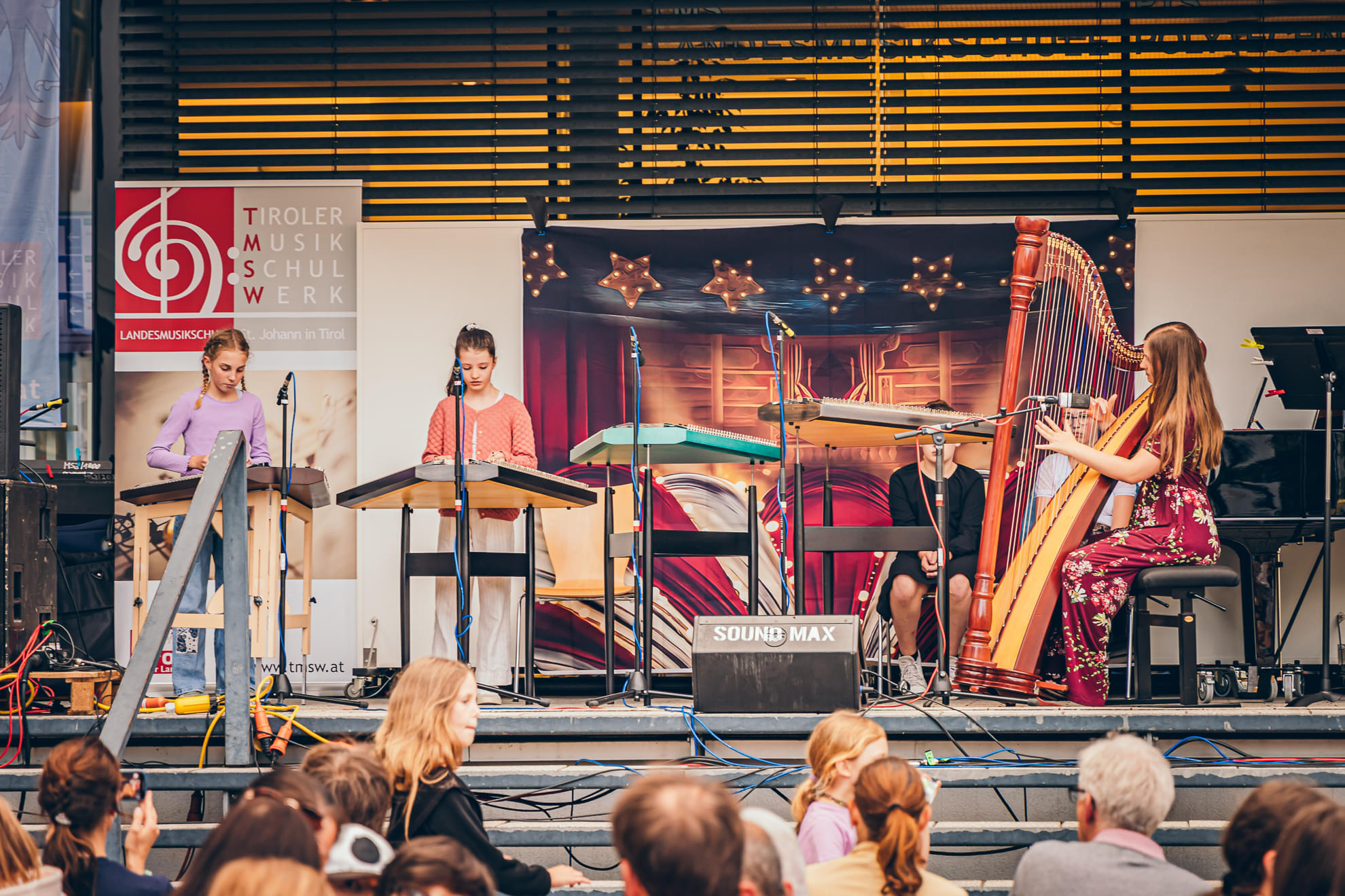 Kinder spielen verschiedene Instrumente auf einer Bühne vor Publikum. Ein Mädchen spielt Harfe, während andere traditionelle Zithern spielen.