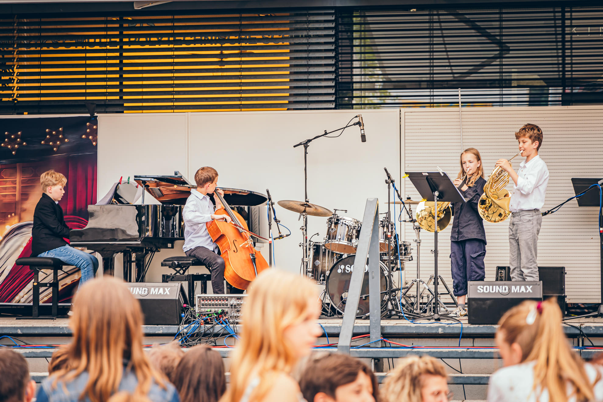 Eine junge Musikgruppe spielt auf einer Bühne mit Klavier, Cello, Schlagzeug und zwei Hörnern. Zuschauer, hauptsächlich Kinder, schauen zu.